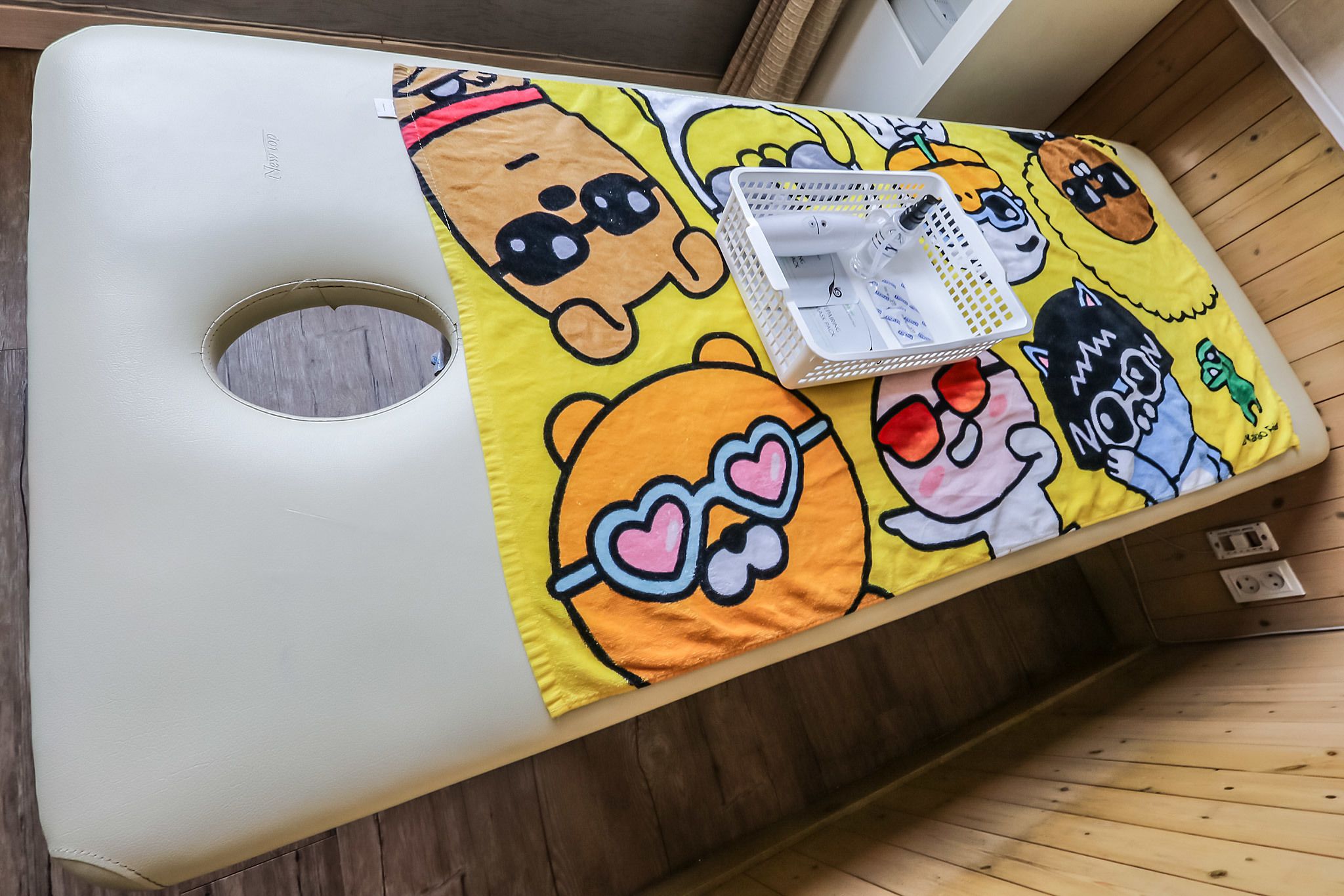 A bed with a colorful blanket featuring cartoon characters, a white pillow, and a white bedside table with a basket containing a toothbrush and toothpaste.