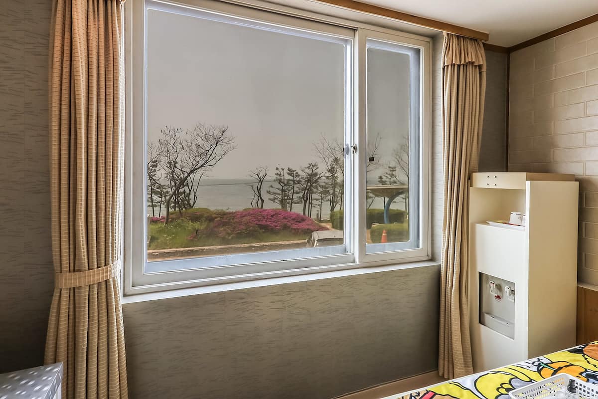 Una habitación con un ventanal grande, cortinas color beige, un armario blanco y una cama con colcha de colores.