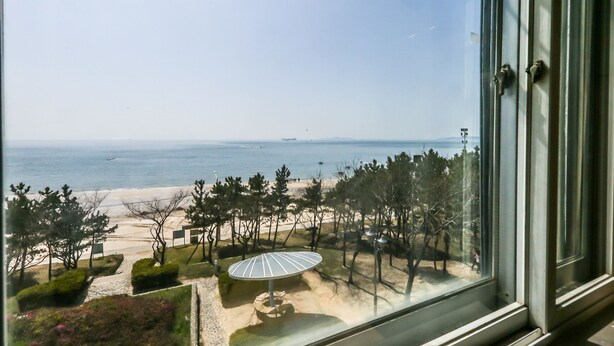 A view from a window overlooking a beach with a sandy area, trees, and a clear sky. - Blue Motel (Boryeong)