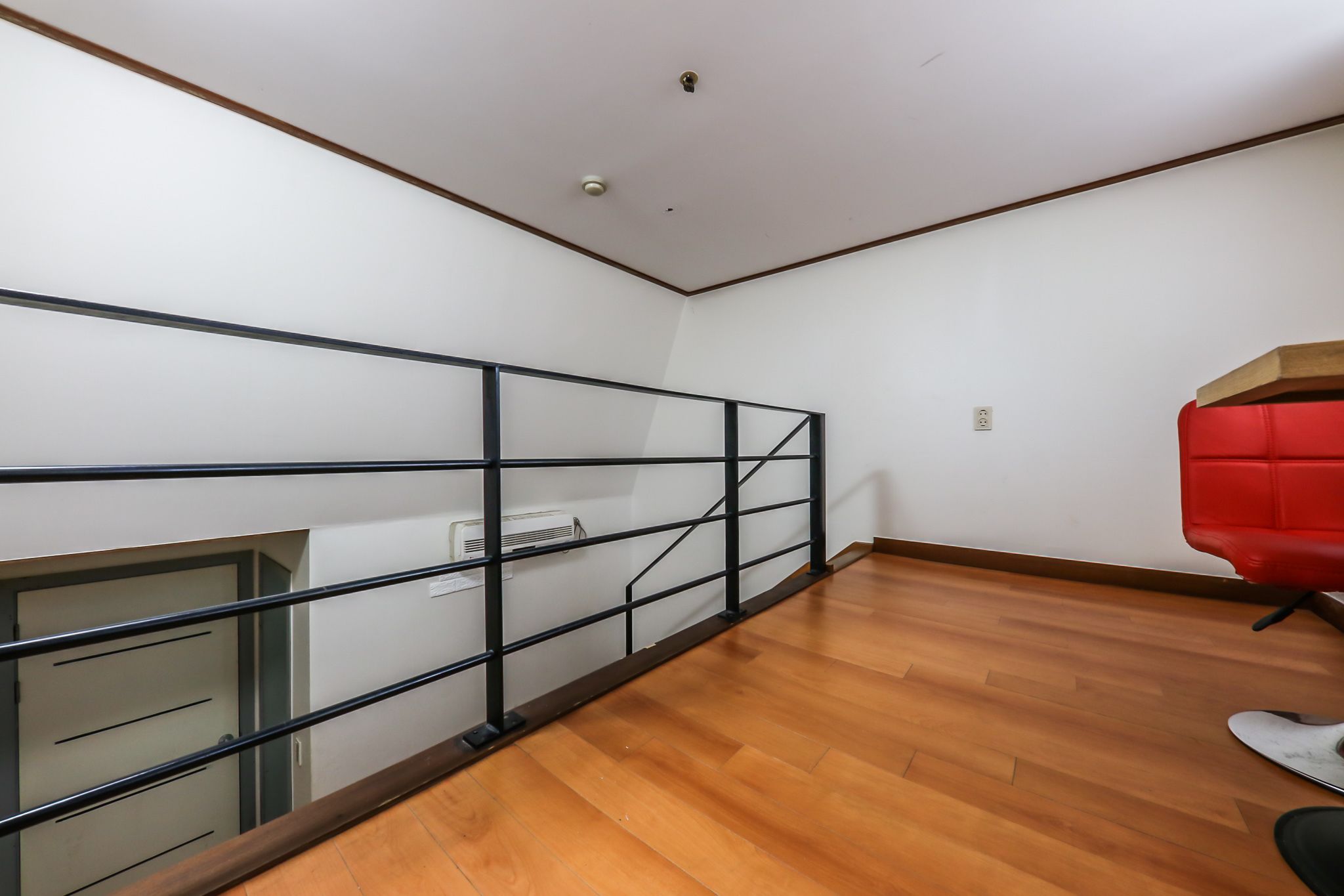A room with a wooden floor, a red chair, a white ceiling, and a black metal railing.
