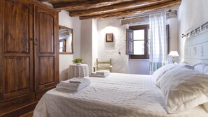Ein Schlafzimmer mit großem Holzschrank, einem Bett mit weißer Bettwäsche, einem kleinen Tisch mit Pflanze und einem Fenster mit Vorhängen.