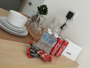 A table with a white bowl, stacked plates, a glass carafe, water bottles, Coca-Cola cans, and Nescafé packets. - The Robertson 1 Bedroom Bukit Bintang KL (Kuala Lumpur)