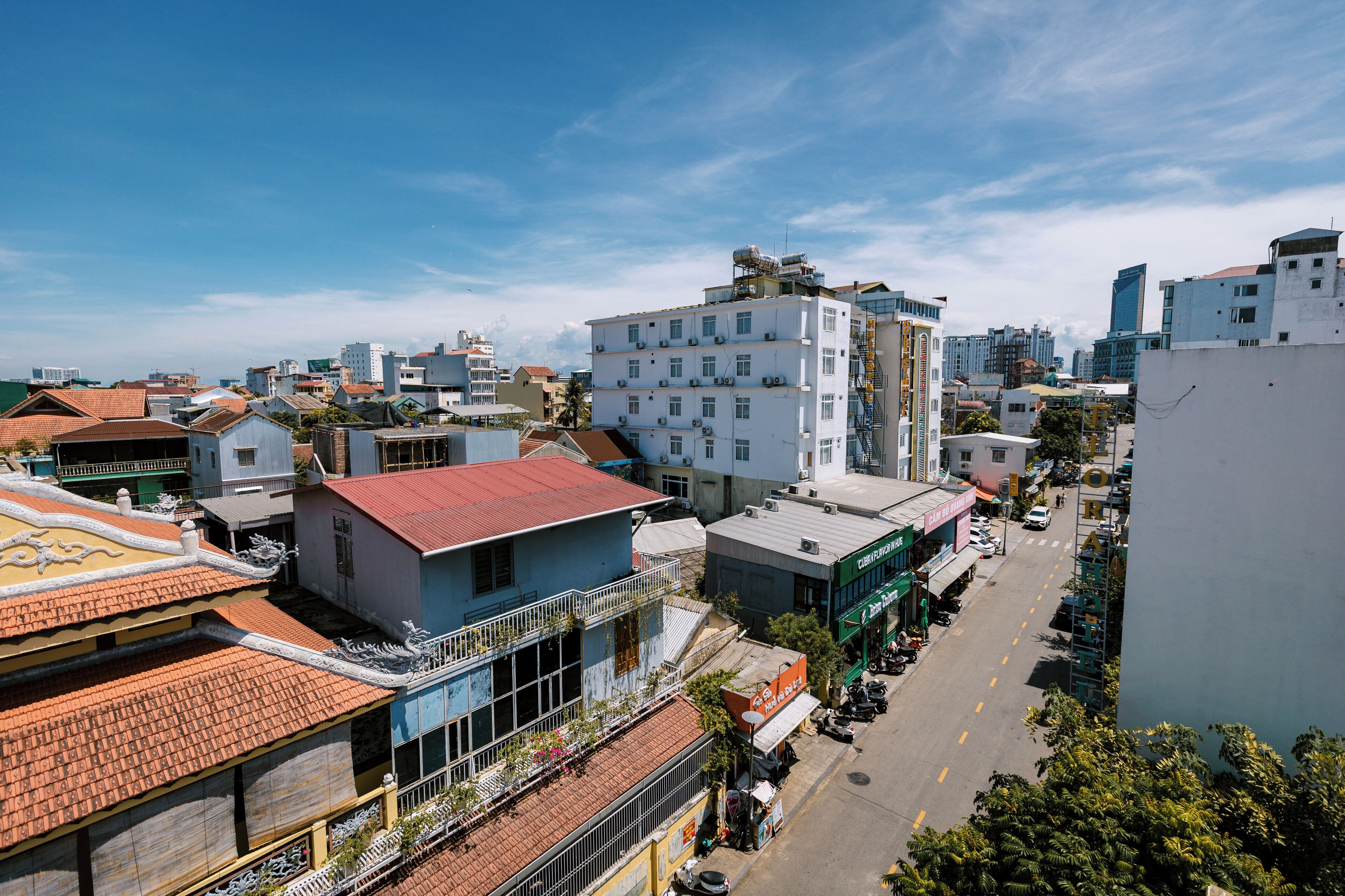 A multi-story building with a red-tiled roof, a balcony with plants, and a street with parked cars.