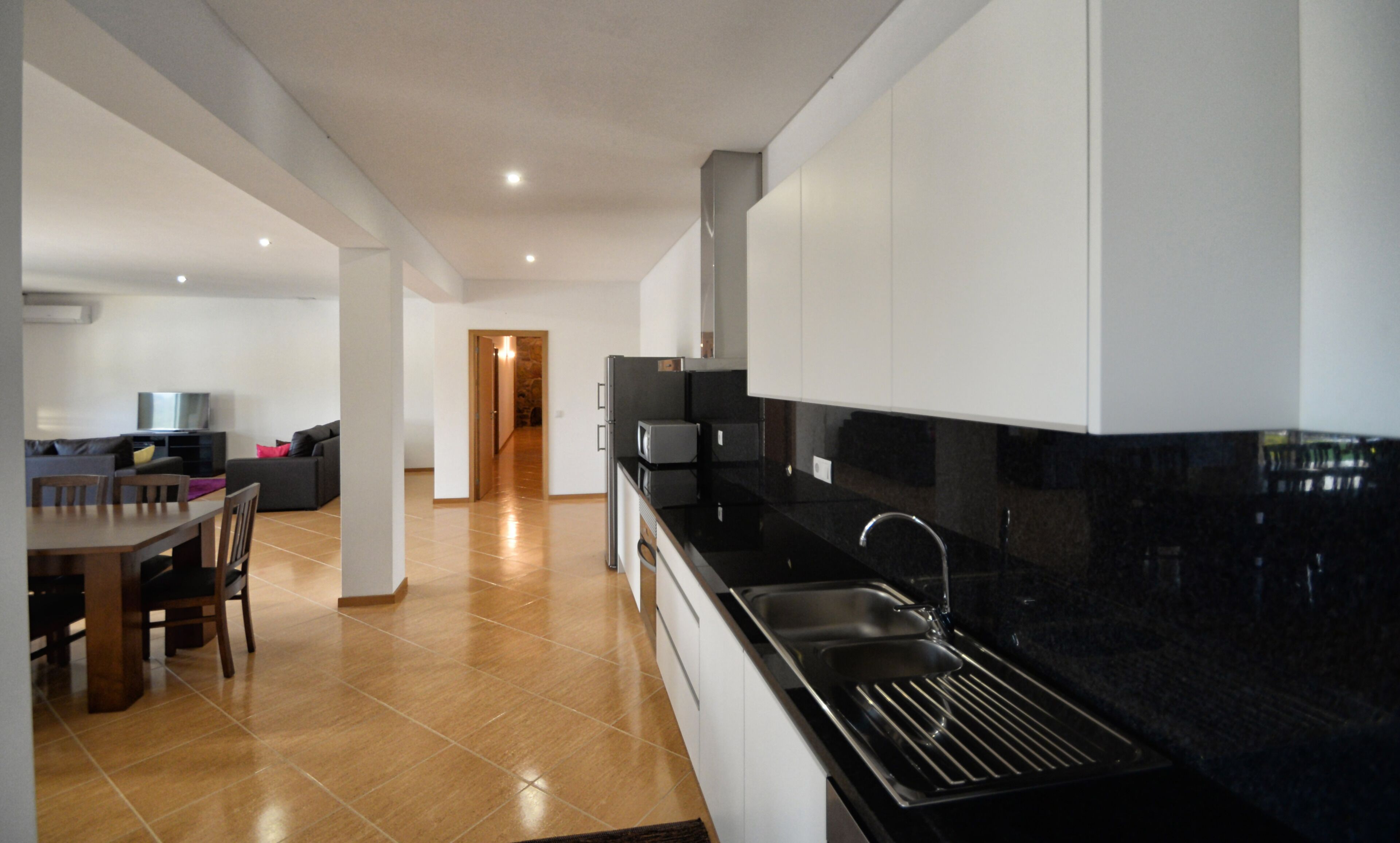 Uma cozinha moderna com bancadas pretas, pia de aço inoxidável e armários brancos. Há uma área de jantar com mesa e cadeiras de madeira, e uma sala de estar com sofá e televisão.