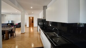 A modern kitchen with black countertops, a stainless steel sink, and white cabinets. There is a dining area with a wooden table and chairs, and a living room with a sofa and a television. - Casa da Caneleira (Camélias de Basto) (Codeçoso)