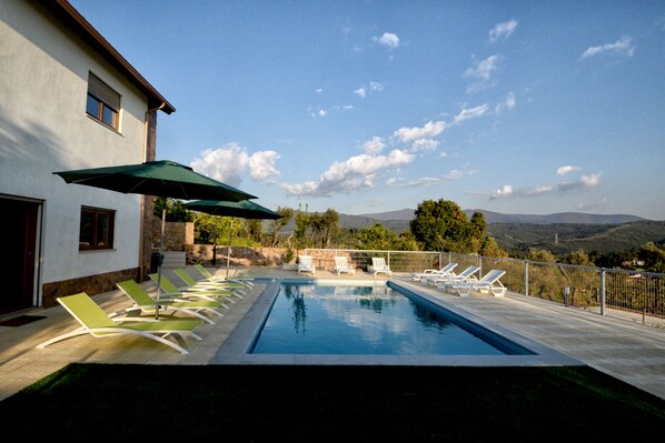 A swimming pool with lounge chairs and umbrellas, a white building, and a mountainous landscape in the background.