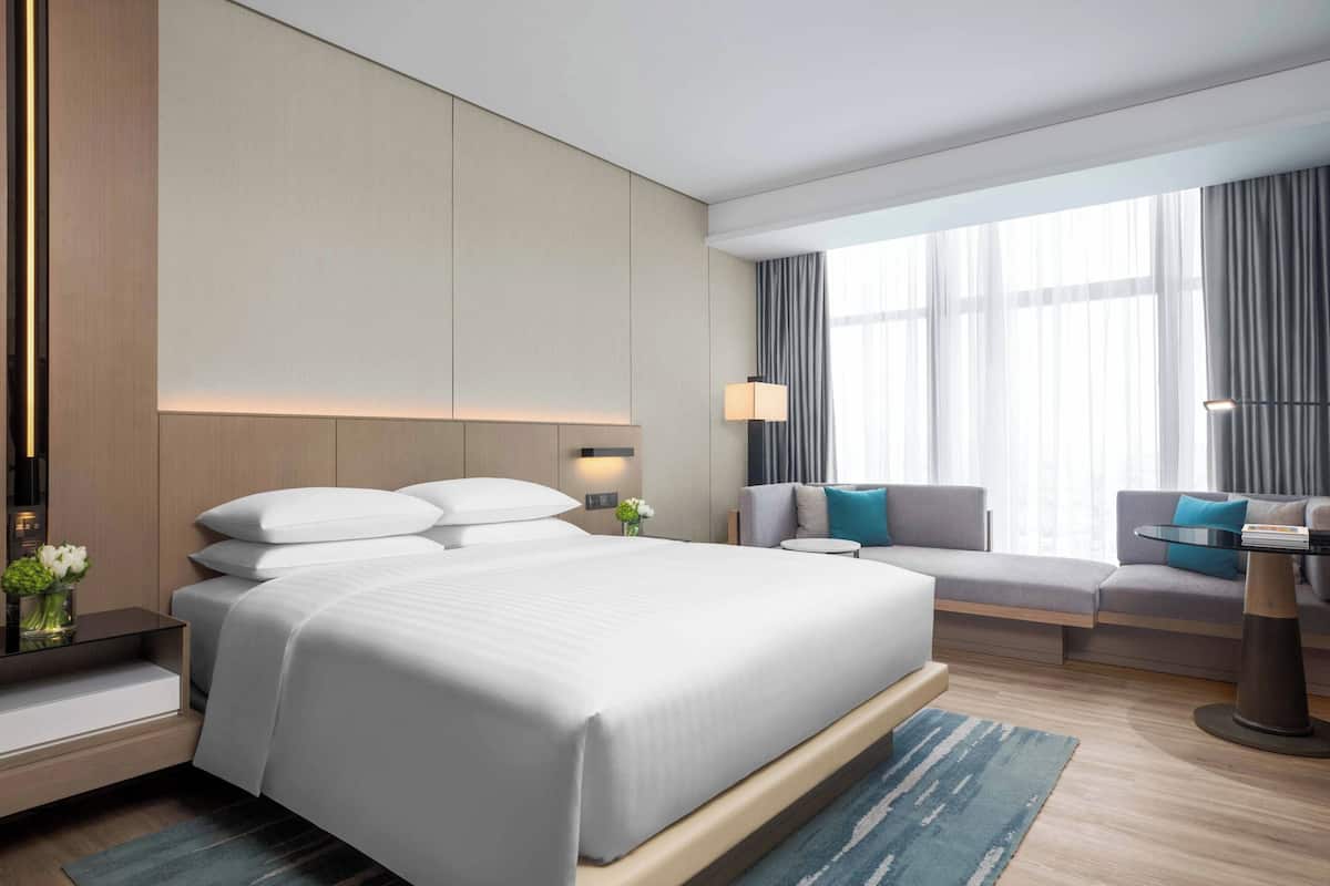 Een moderne hotelkamer met een groot bed, een bank, een bureau en uitzicht op de stad door grote ramen.
