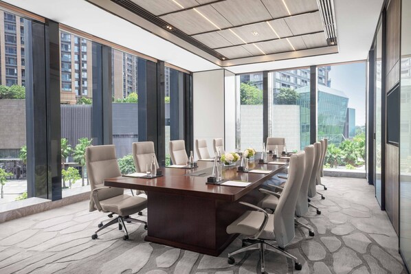 Un salón de conferencias con una gran mesa, varias sillas y vistas a edificios y espacios verdes al exterior.