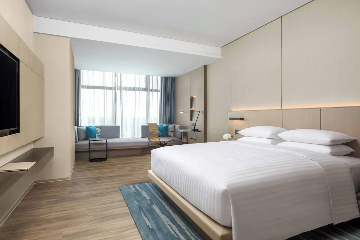 Een moderne hotelkamer met een groot bed, een flatscreen televisie aan de muur, een zithoek met een bank en fauteuils, en een groot raam dat voor natuurlijk licht zorgt.