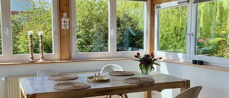 Una mesa de comedor de madera con sillas, un florero con flores y vista a la vegetación a través de amplios ventanales.