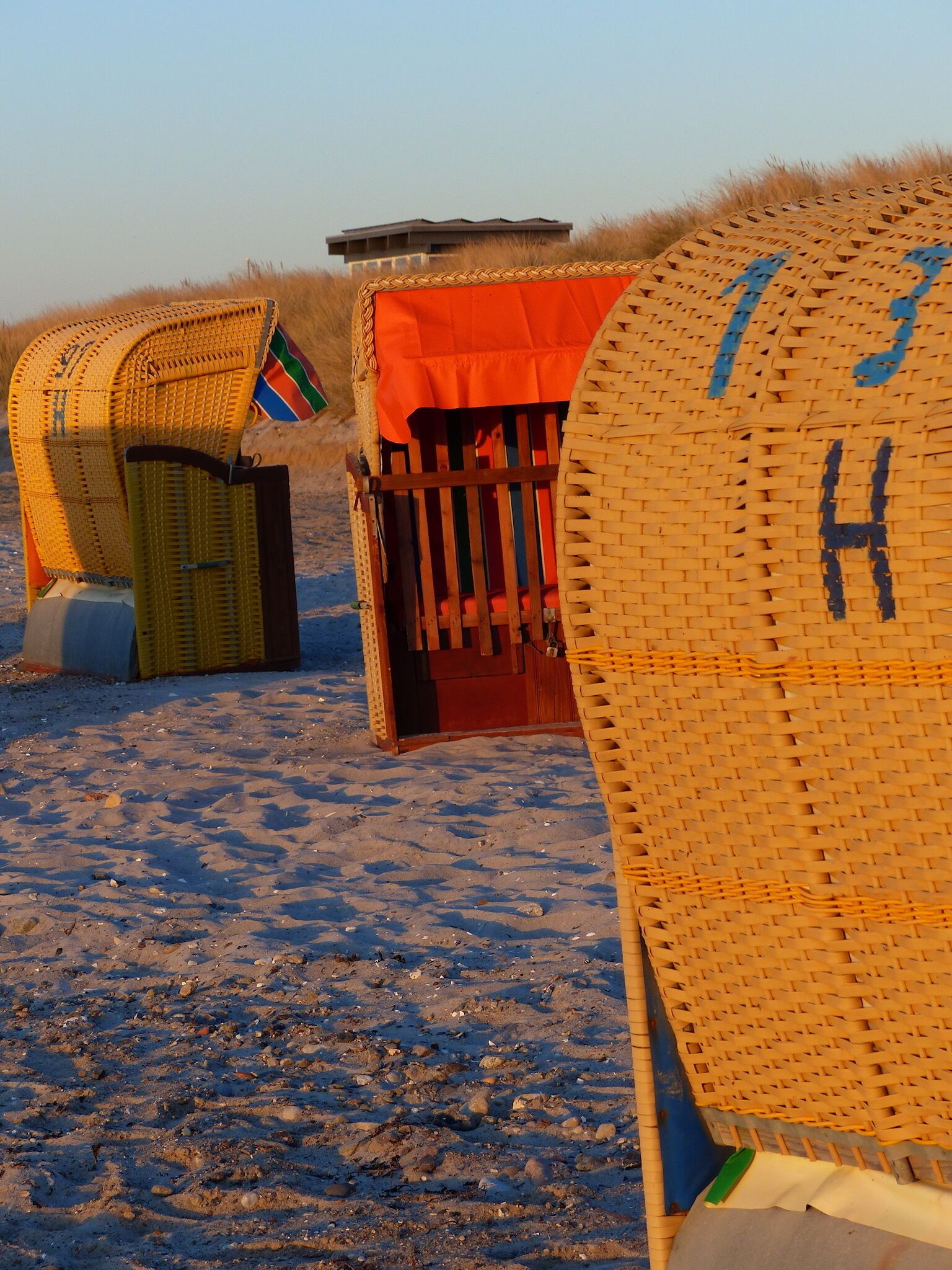Een strandscène met een grote rieten mand met de tekst 'H' en een klein rood gebouw.