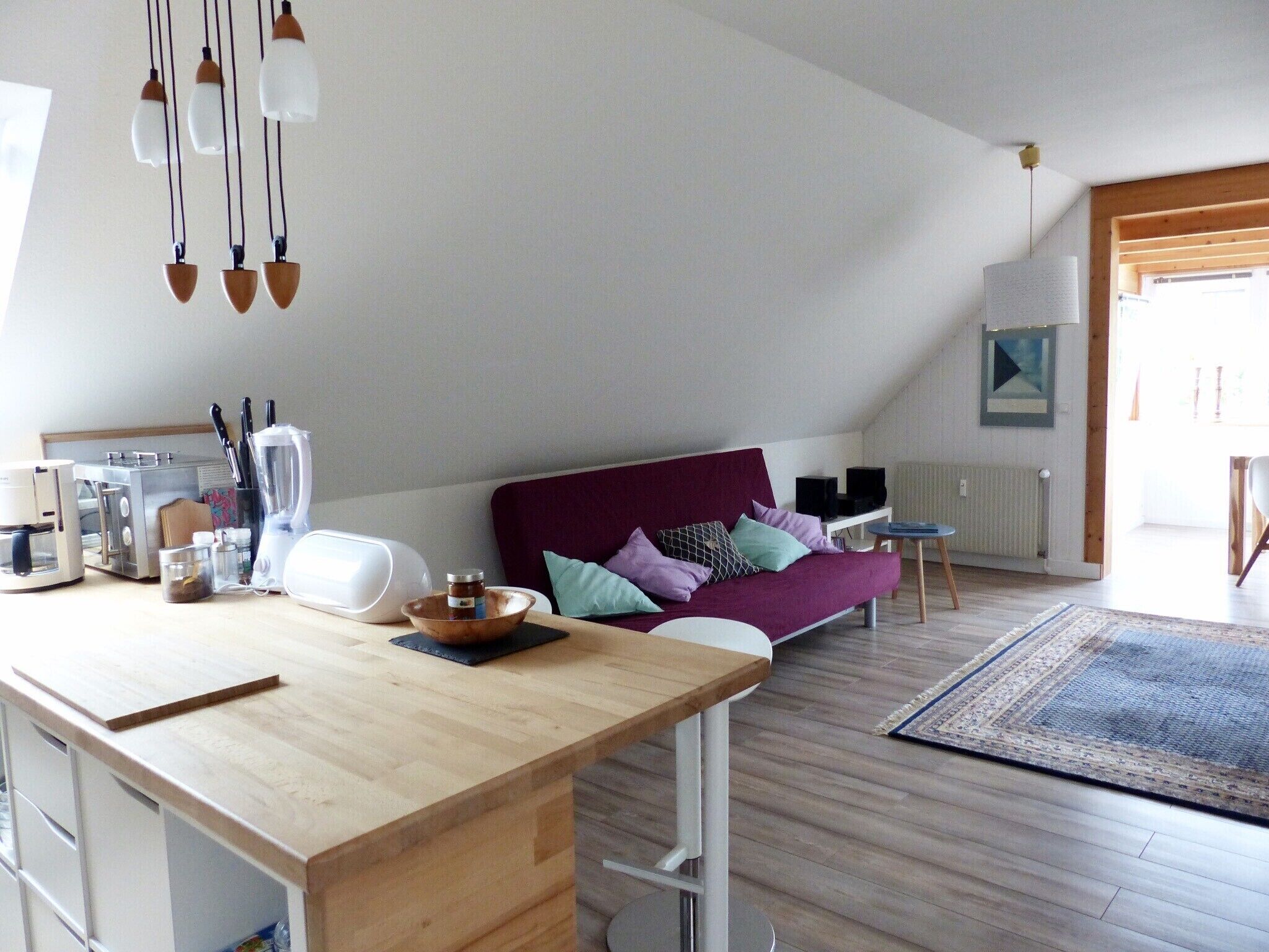 Una cocina moderna con encimera de madera, zona de comedor con sofá púrpura y vistas al exterior a través de un ventanal.