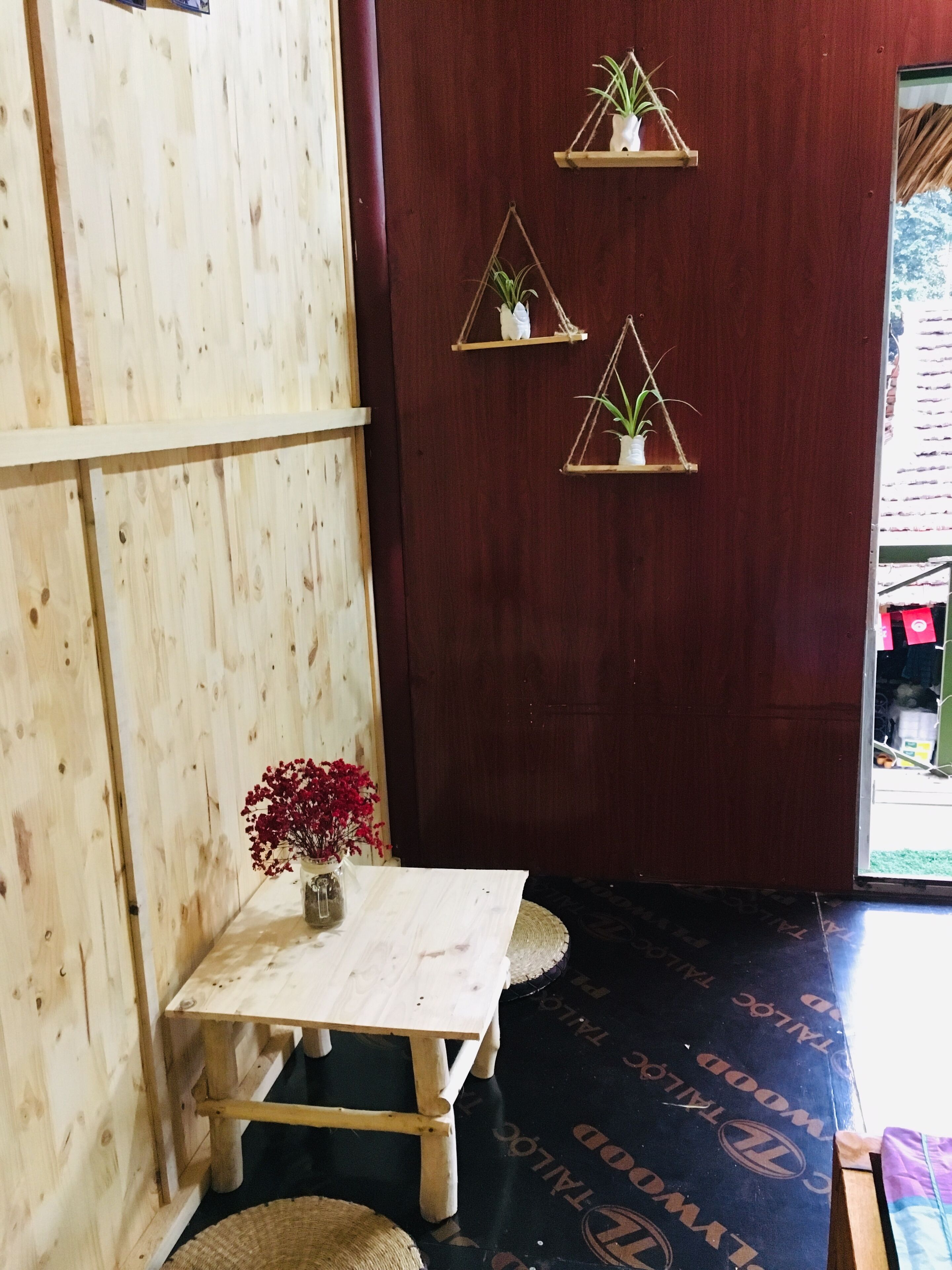 A wooden room with a small table and a vase of red flowers. There are three hanging plant shelves on the wall.