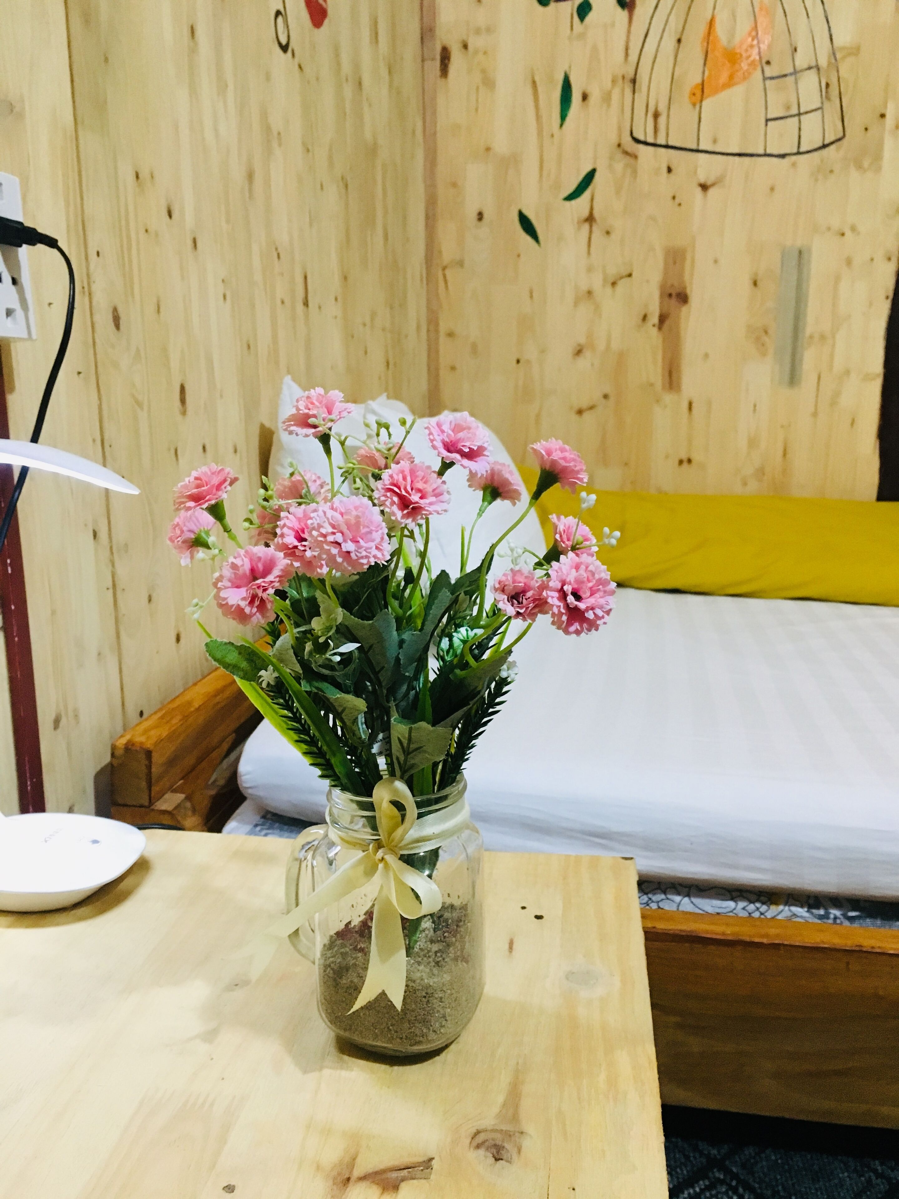 A wooden room with a bed, a small table with a vase of pink flowers, and a wall with a birdcage design.