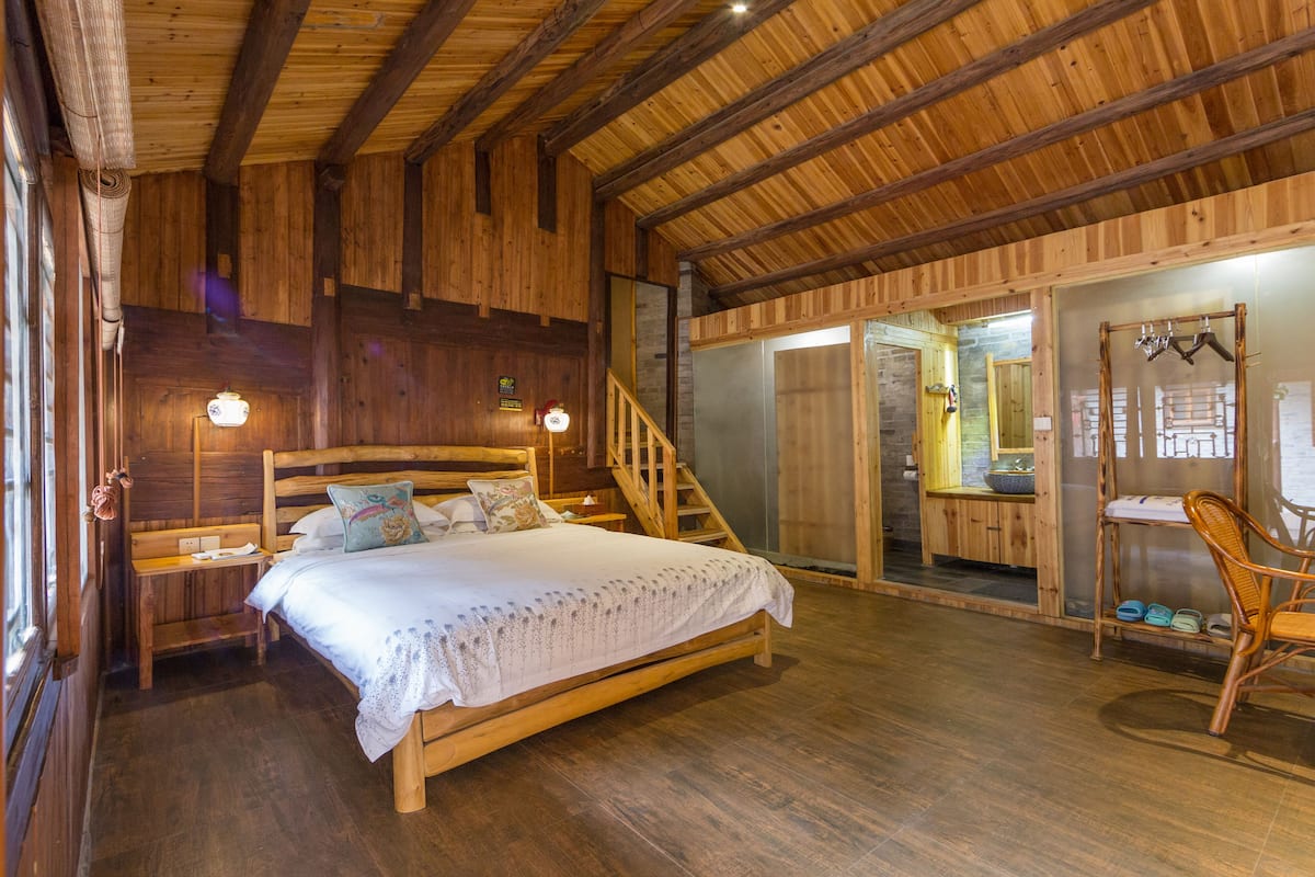 Ein Schlafzimmer einer Holzhütte mit einem Bett, einem Sessel, einer Treppe und einem durch eine Glastür einsehbaren Badezimmer.