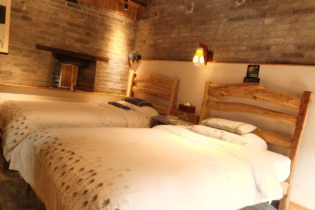 Ein rustikaler Raum mit zwei Holzkohlenbetten, einer Backsteinwand und einem kleinen Tisch mit Lampe.