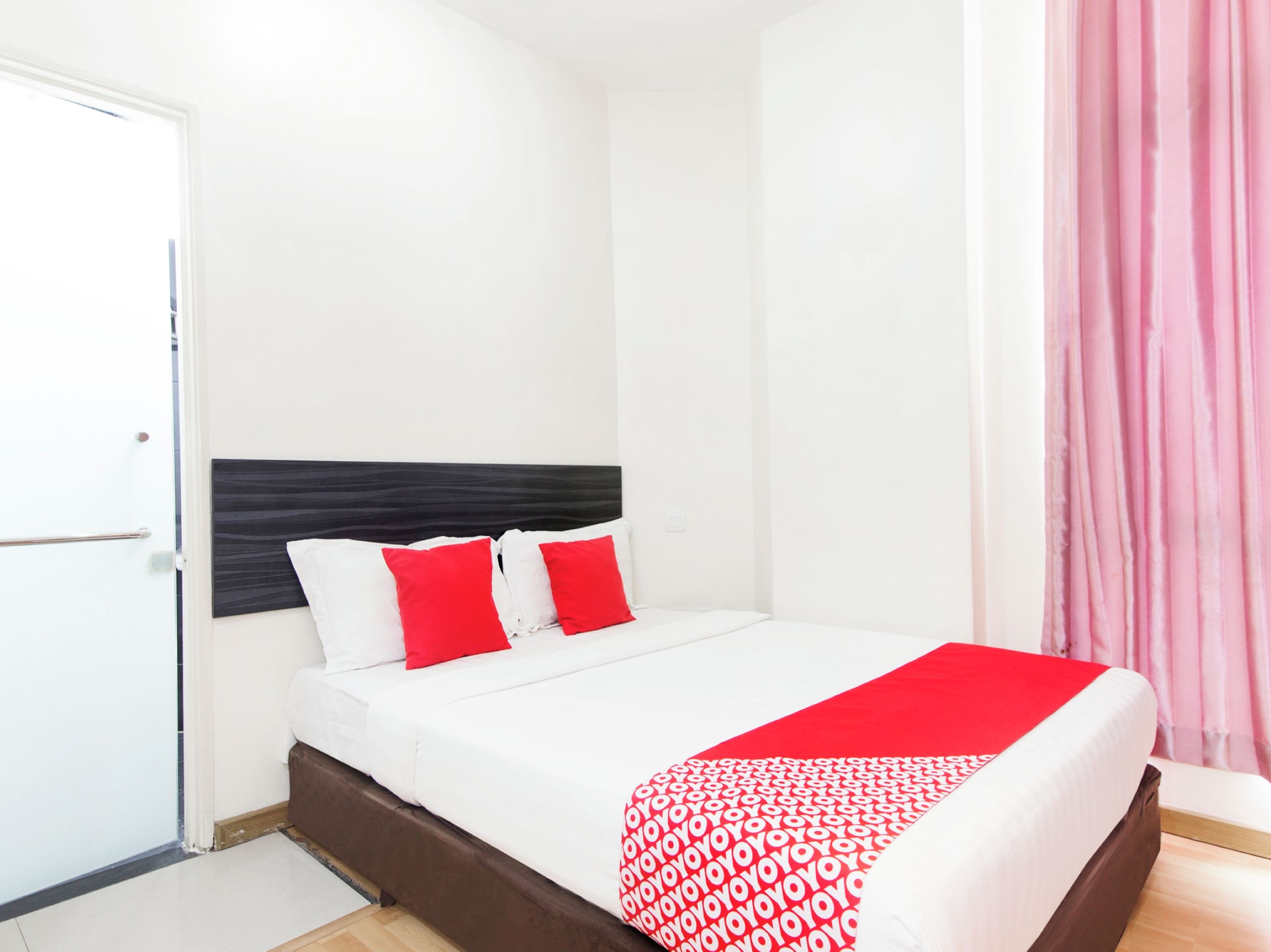 A neatly made bed with white and red bedding, a dark headboard, and a pink curtain on the window.