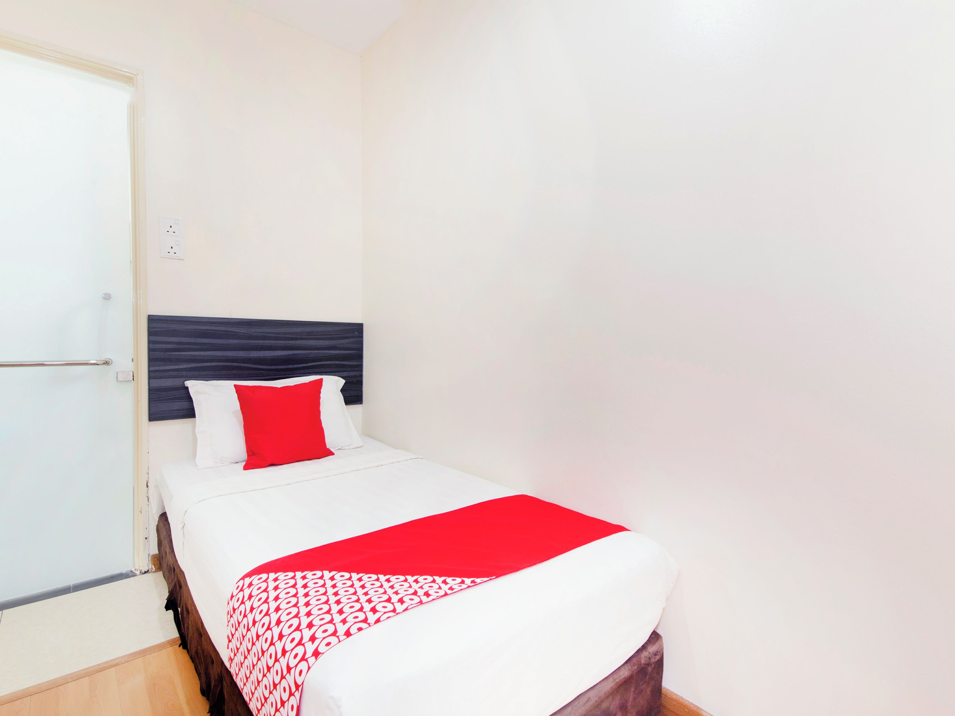 A small, neatly made bed with a white and red bedspread, a dark headboard, and a white pillow.