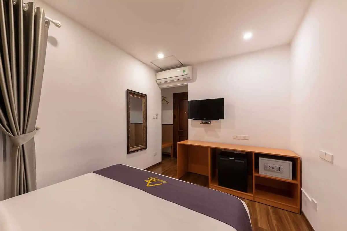 A hotel room with a bed, a TV, a small wooden cabinet, and an air conditioning unit.