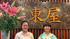 Two receptionists at a hotel reception desk with a sign that reads "東屋". - Azumaya Hotel Linh Lang (Hanoi)