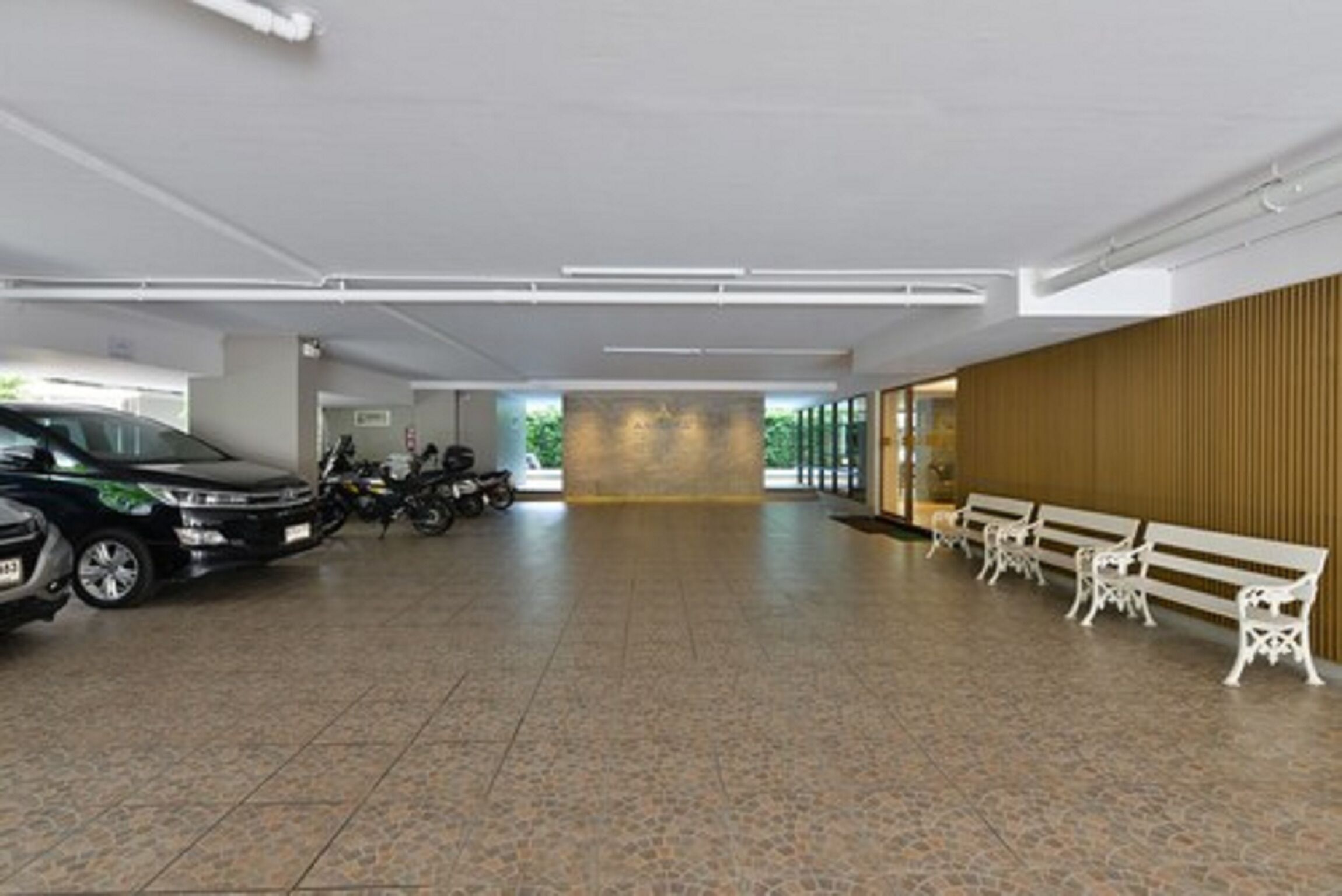 A spacious indoor area with tiled flooring, a row of white benches, and a car and motorcycle parked in the background.