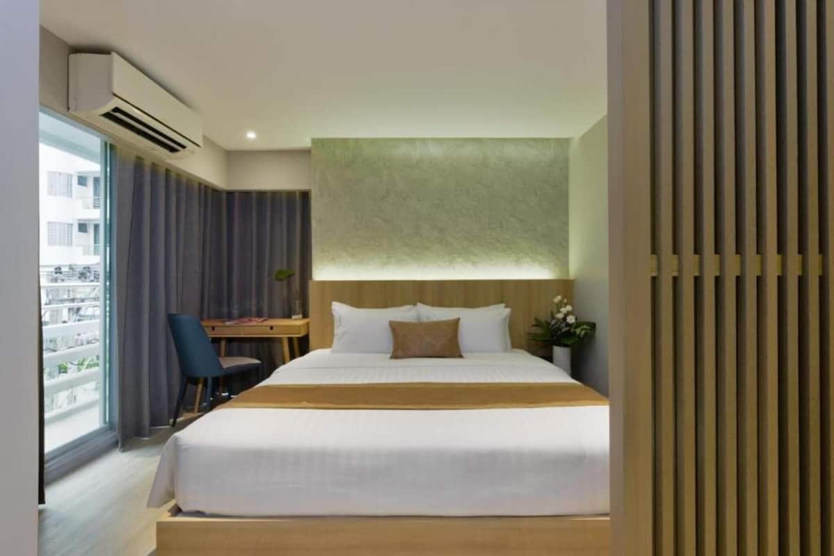 A hotel room with a bed, a desk, a chair, a window with curtains, and a view of buildings outside.