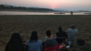 En gruppe mennesker sidder på en sandstrand ved solnedgang, med en flod i baggrunden.