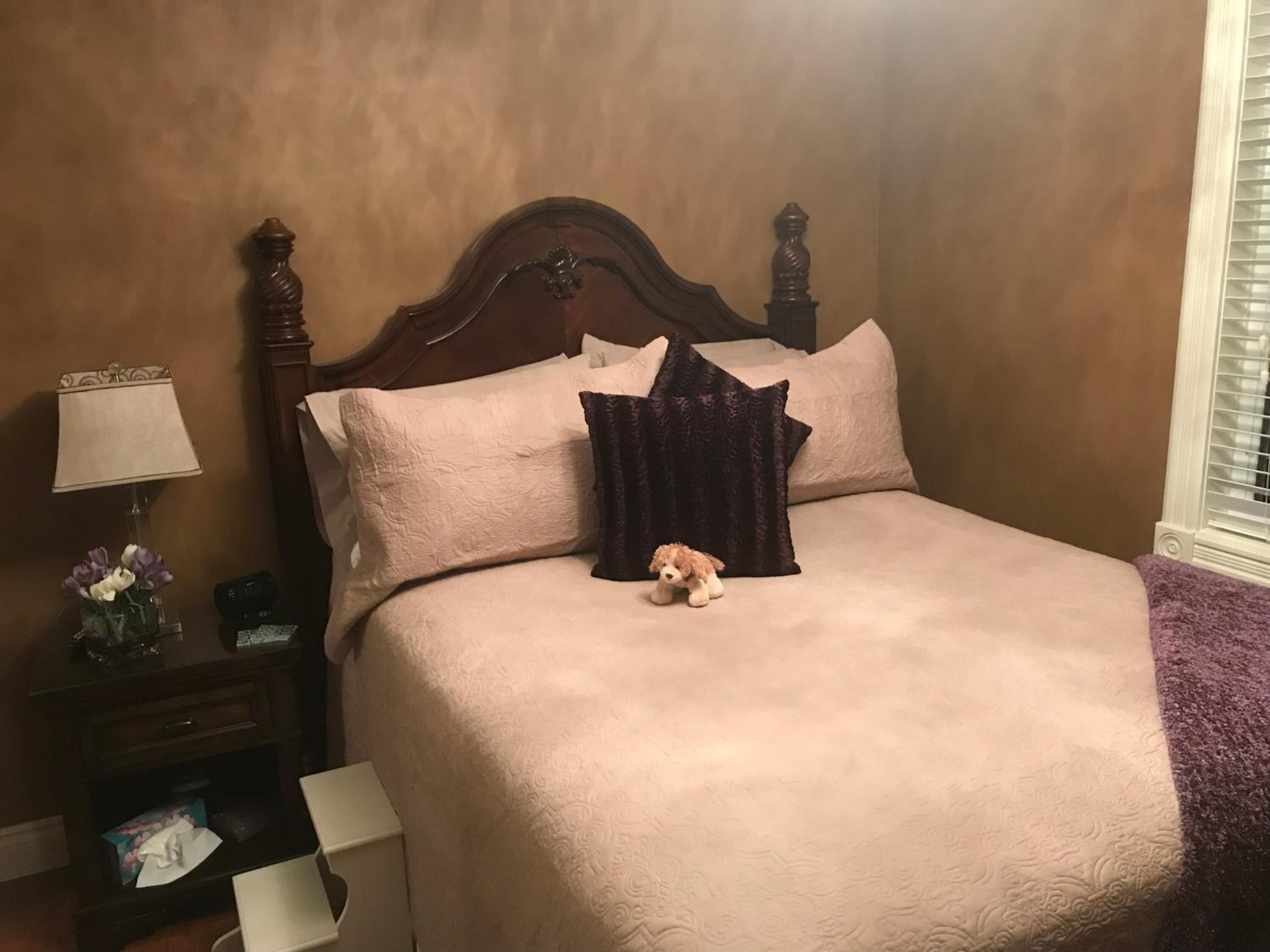 A bedroom with a wooden headboard, a bed with beige bedding, a small nightstand with a lamp and a vase of flowers, and a window with blinds.