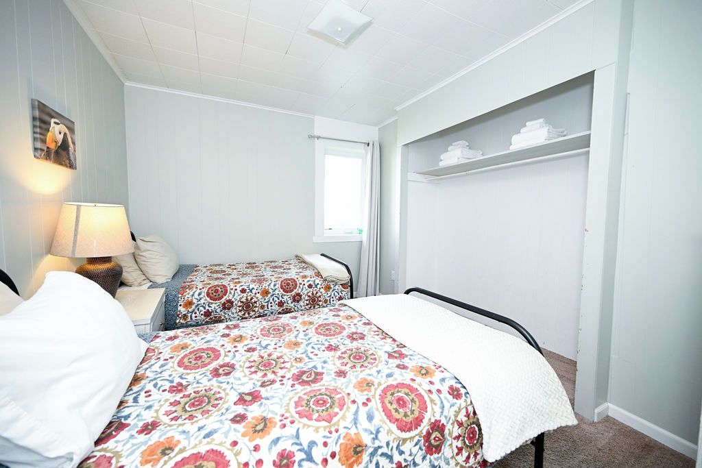 A bedroom with two beds, a nightstand, a lamp, a window, and a built-in shelf with folded towels.
