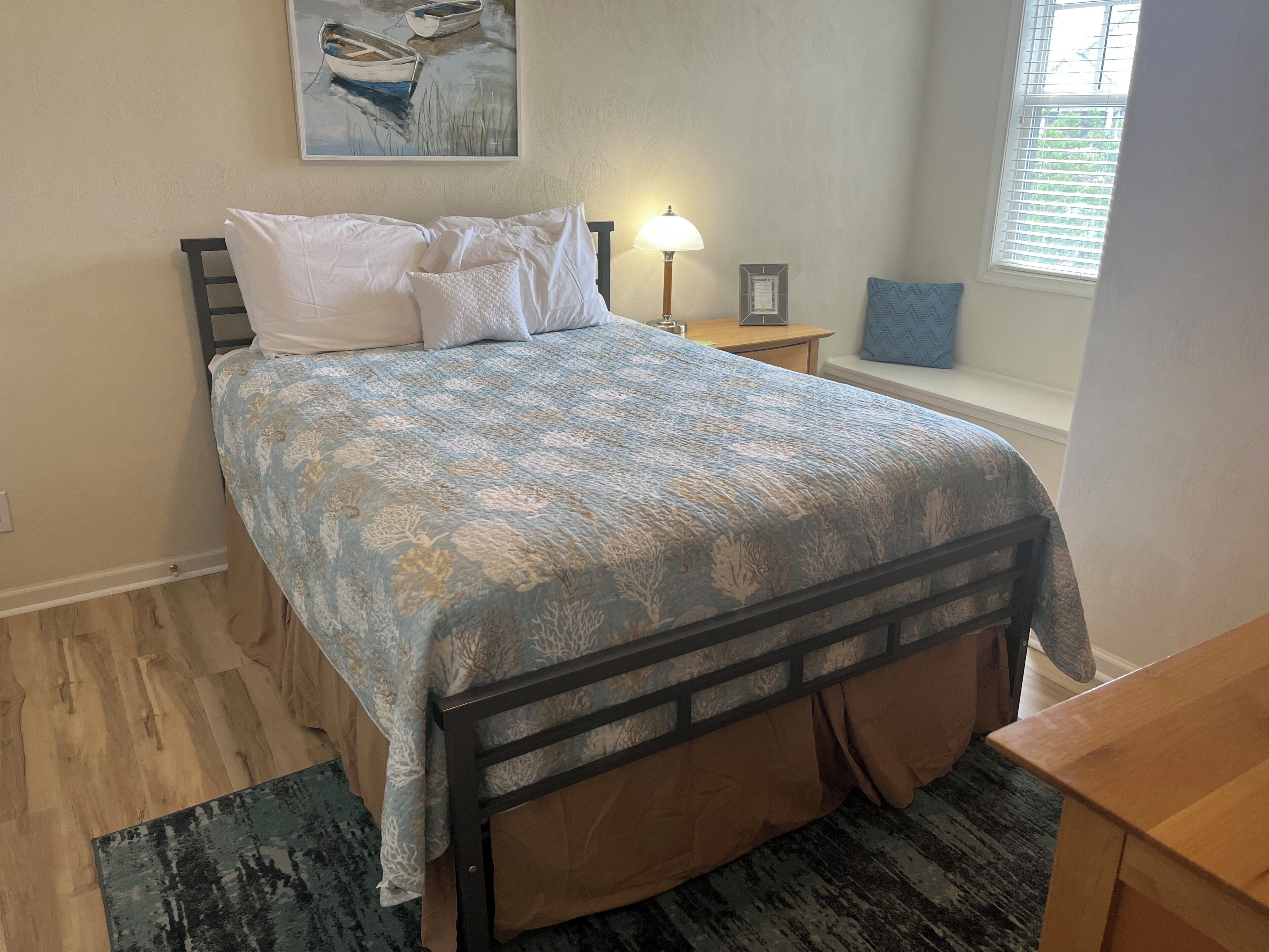 Una cama bien hecha con una colcha estampada, una lámpara de noche, un cuadro en la pared y una ventana con persianas.