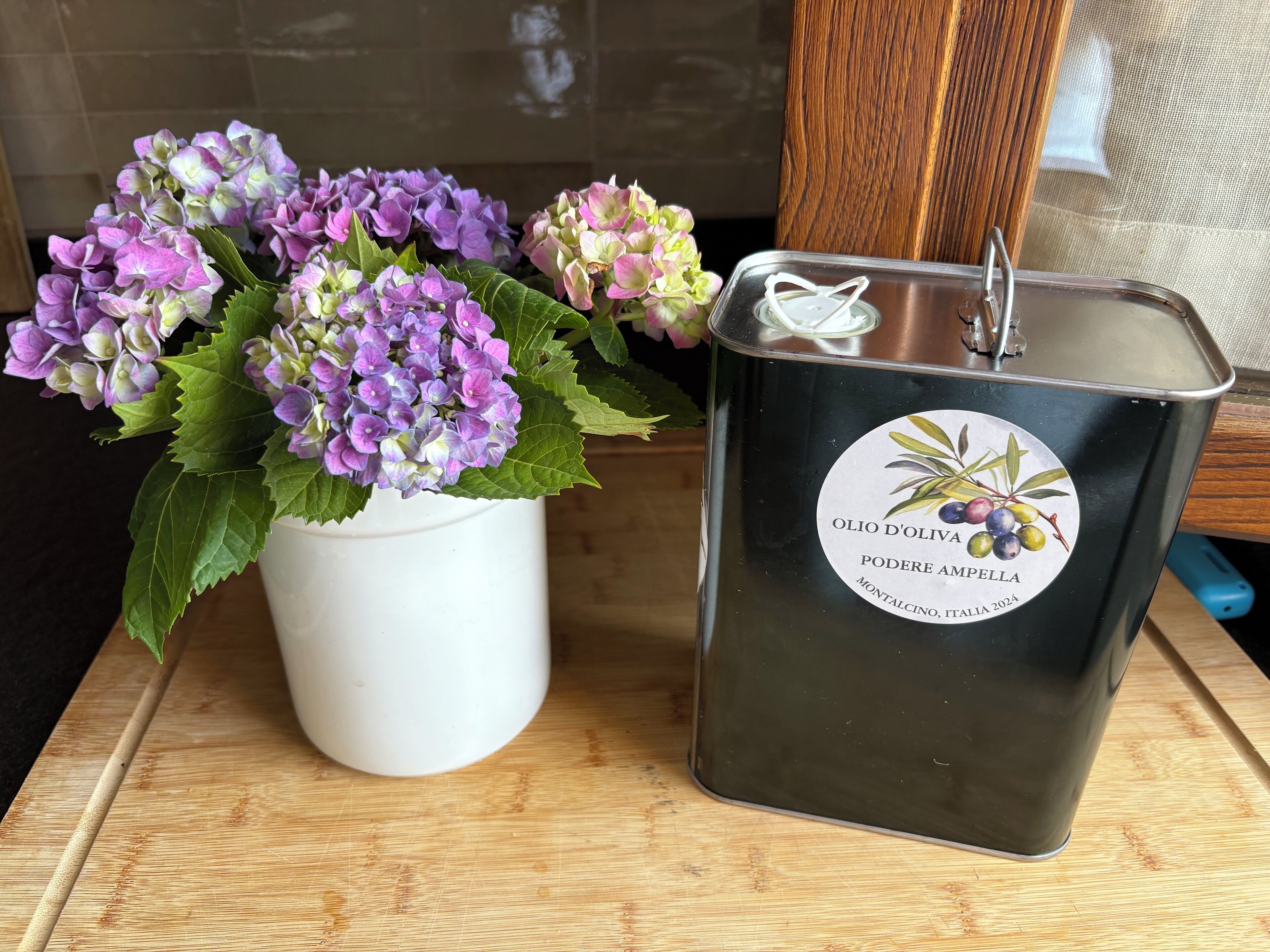 Una maceta blanca con flores de hortensia moradas y una vela negra con etiqueta.