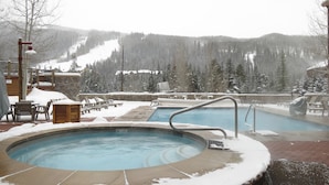 A hot tub area with a pool, surrounded by snow-covered chairs and tables, with a view of a snowy mountain landscape.