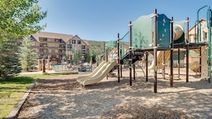 A playground with slides and climbing structures, surrounded by a wooded area and a building with multiple windows and balconies.