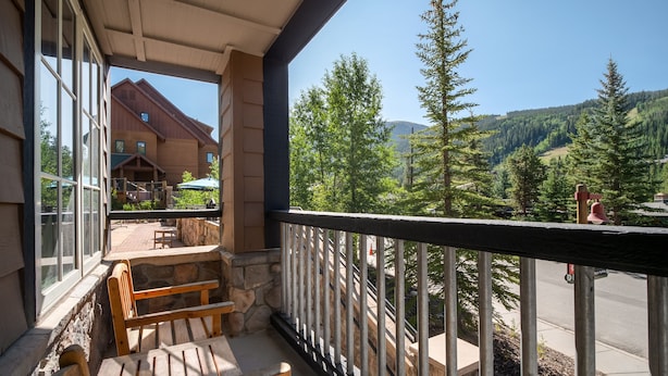 A balcony with wooden furniture, a stone wall, and a view of a lodge and forested mountains.
