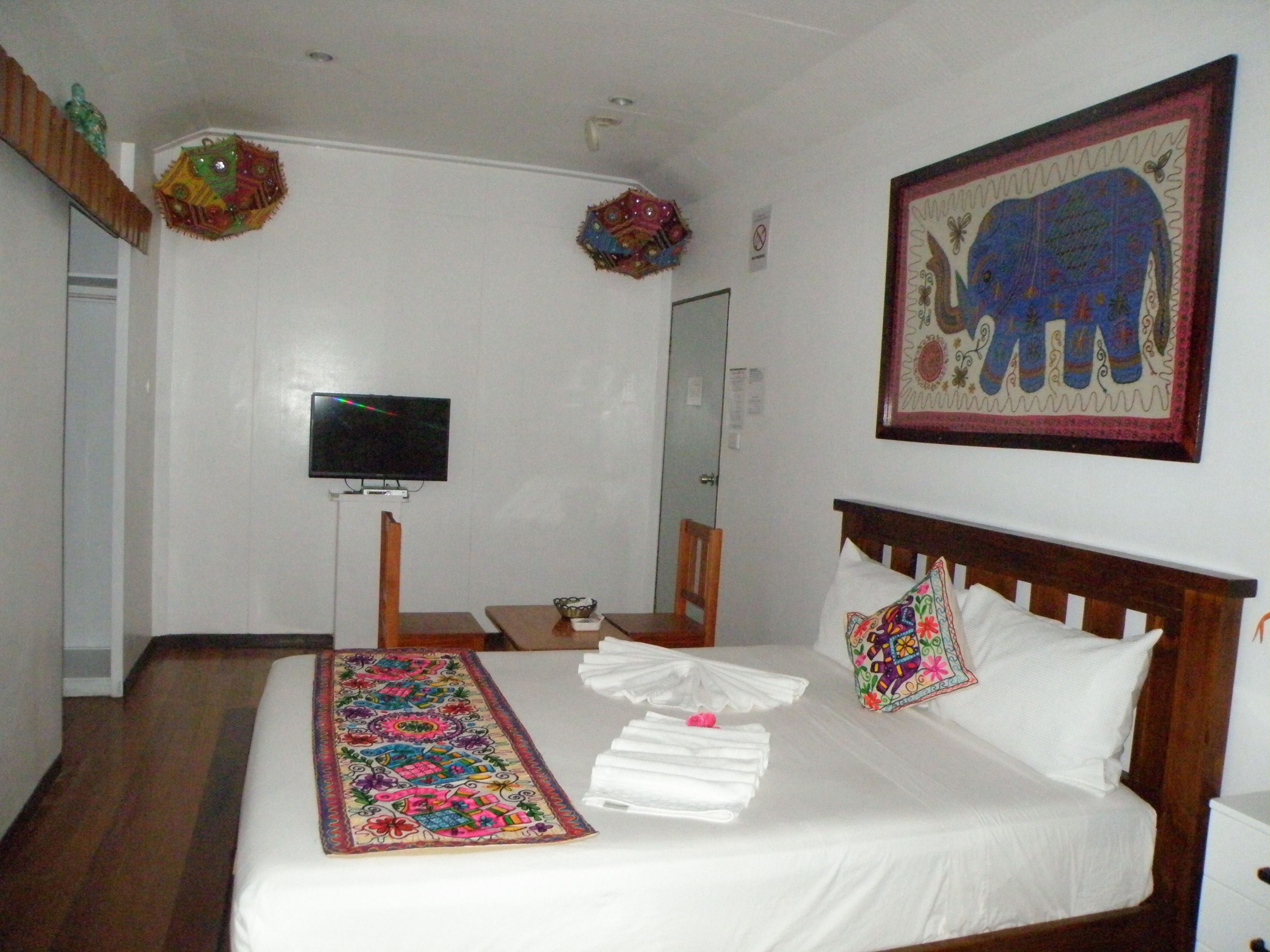 A hotel room with a bed, a TV, a wooden headboard, a colorful artwork, and a decorative wall hanging.