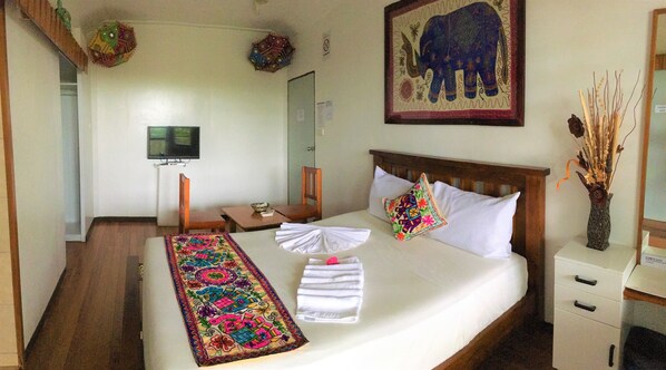A neatly made bed with a colorful runner, a wooden headboard, two pillows, a small table with a chair, a TV on a stand, and a framed elephant painting on the wall. - Tagimoucia House Hotel (Suva)