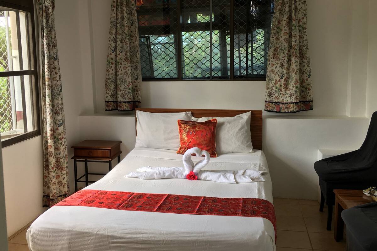 A neatly made bed with a red and white patterned bedspread, a heart-shaped towel decoration, and a wooden nightstand beside it.