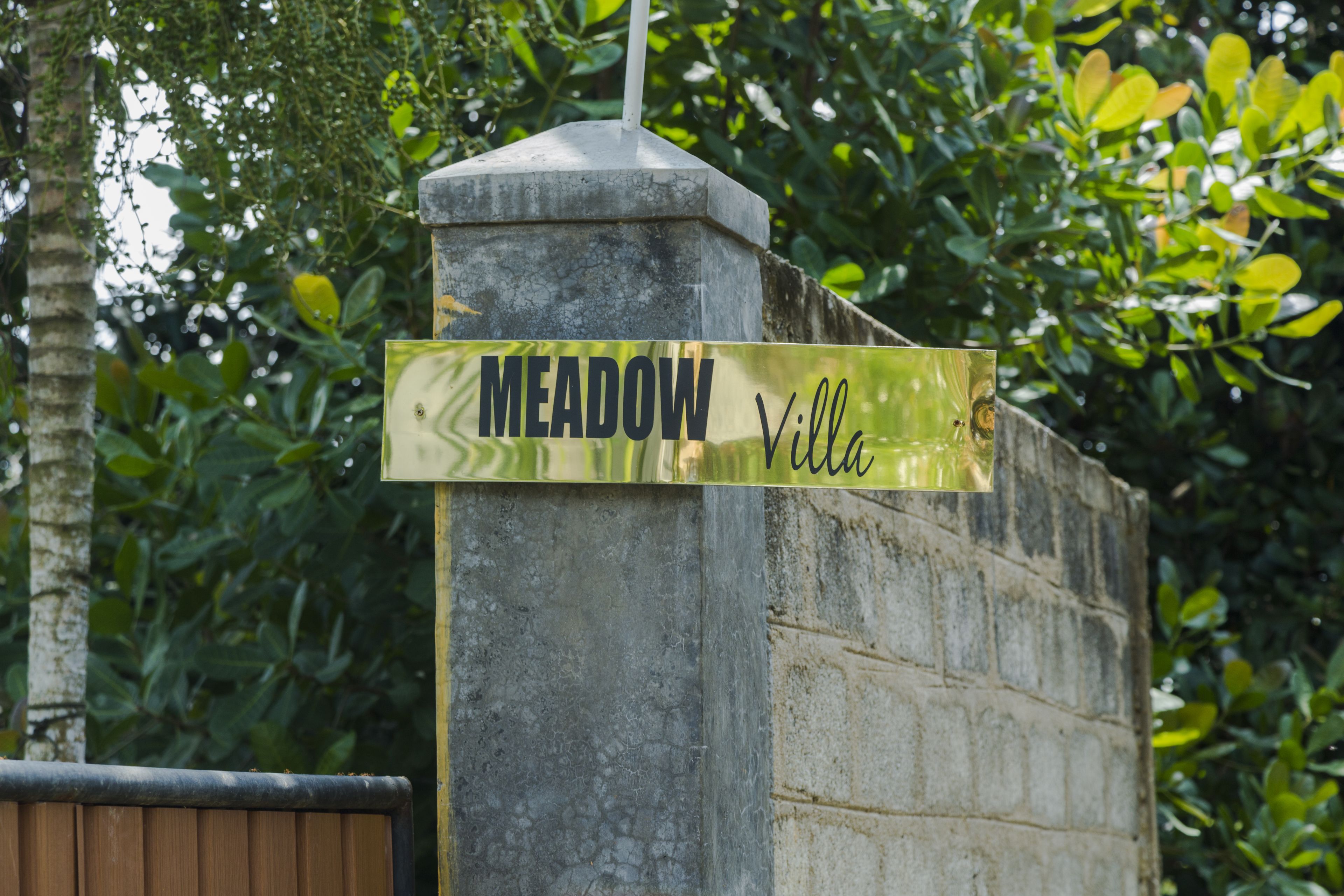 En skylt med texten ”MEADOW Villa” är fäst på en stenkantad pelare, med en mur av tegelstenar och grönska i bakgrunden.