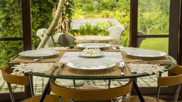 A dining table set for four with white plates, silverware, and a centerpiece of white flowers. The table is surrounded by chairs and set against a backdrop of lush greenery and a house. - Meadow Villa - Galle (Galle)