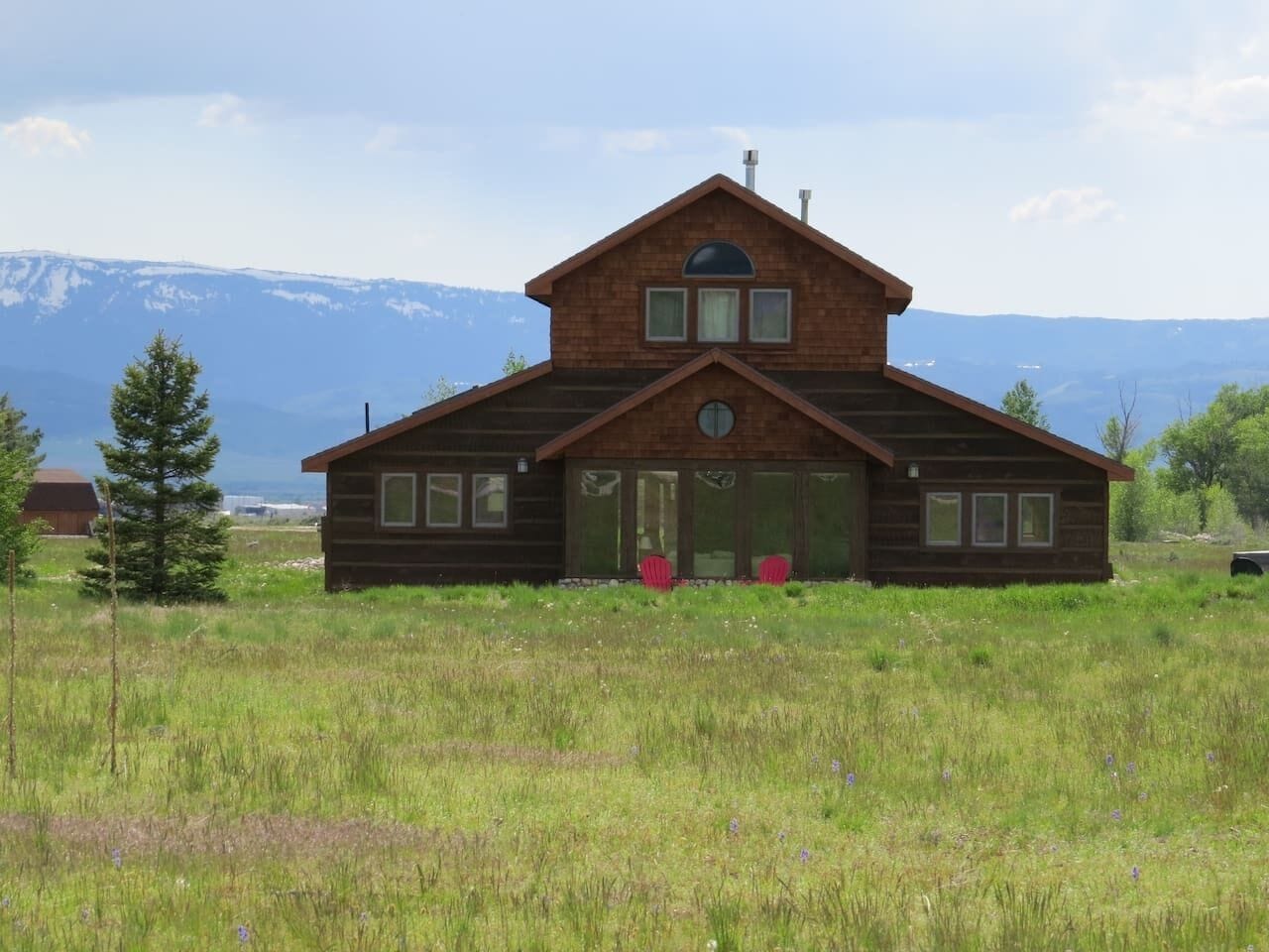 A wooden cabin with a gabled roof, large windows, and a door with a glass panel, surrounded by a grassy field and mountains in the background.