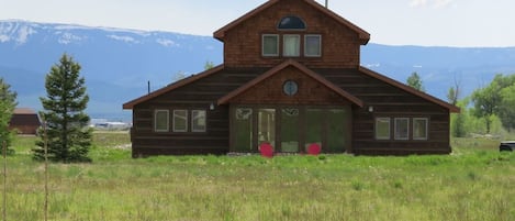 A wooden cabin with a gabled roof, large windows, and a door with a glass panel, surrounded by a grassy field and mountains in the background.