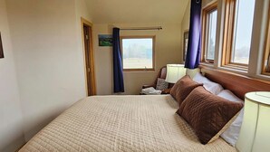 Un dormitorio con una cama grande, una mesita de noche con lámpara, una ventana con cortinas y una puerta que da a otra habitación.