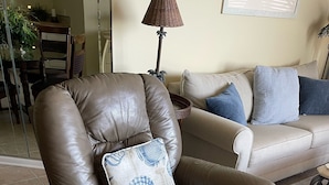 Ein brauner Leder-Sessel mit dekorativem Kissen, ein beiges Sofa mit blauen Kissen und eine Stehlampe in einem Wohnzimmer mit Fliesenboden.