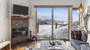 A living room with a stone fireplace, a flat-screen TV, and a large window with a snowy mountain view. - Outpost: Harvest Dance 3150 - Teton Views (Jackson)