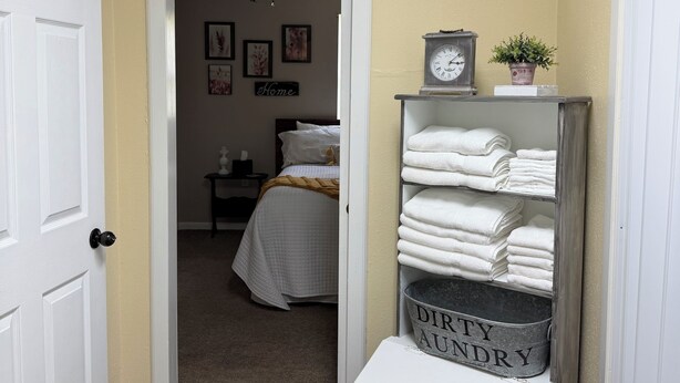 Un pasillo que conduce a un dormitorio con cama, una mesita de noche, un reloj de pared y un estante con toallas dobladas.
