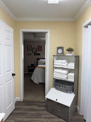 Un pasillo que conduce a un dormitorio con cama, una mesita de noche, un reloj de pared y un estante con toallas dobladas.
