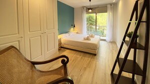 A bedroom with a bed, a chair, a bedside table, a window with curtains, and a wall-mounted shelf.
