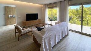 A modern living room with a large flat-screen TV, wooden furniture, and a balcony with a view of greenery.