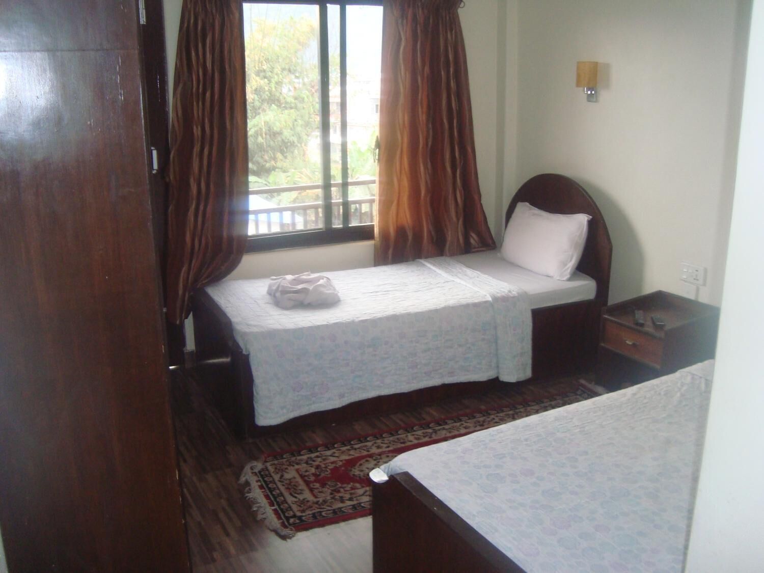 Una habitación pequeña y sencilla con una cama individual, una mesita de noche, una ventana con cortinas y una lámpara fijada a la pared.
