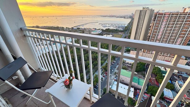 Un balcon avec une table et des chaises, donnant sur un paysage urbain composé d’immeubles et d’une étendue d’eau, au moment du coucher du soleil.